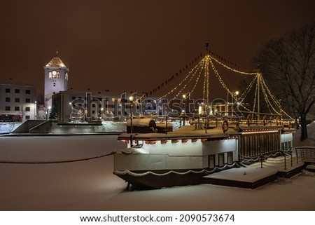 Colorful ship with festive lights frozen in river at Christmas time. Restaurant on water at winter. Pedestrian bridge across the river  in water. Winter landscape with cafeteria in shape of boat. 
