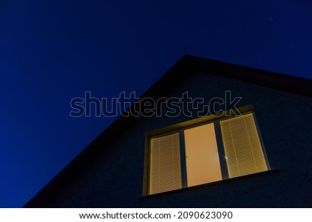 House roof and window with lights on against a blue night sky. Web banner.