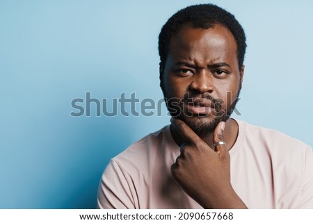 Black bristle man in t-shirt frowning while holding his chin isolated over blue background
