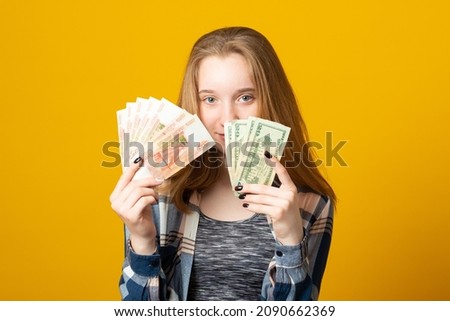 Young woman in casual clothes holds money, dollars in one hand and Russian rubles in the other on a yellow background.	
