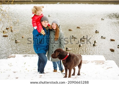 Full height family portrait with one kid and dog in winter casual outfit posing outdoors near river with duck birds, weekend activities and fresh air