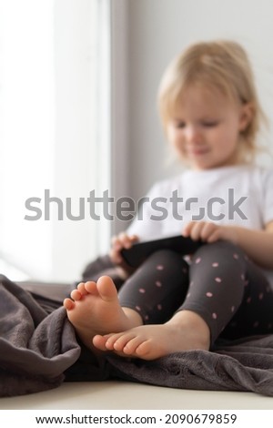 Blurred image of little girl with blond hair sitting on the window wrapped in blanket and using mobile device.
