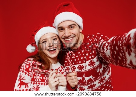 Close up fun young couple friends two man woman 20s in sweater hat do selfie shot pov on mobile phone hold candy cane lollipop isolated on plain red background Happy New Year 2022 celebration concept