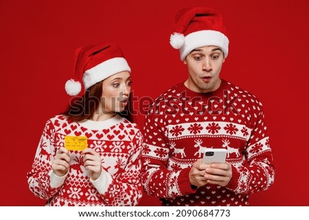 Young amazed couple friends two man woman in sweater hat hold credit bank card do online shopping hug isolated on plain red background Happy New Year 2022 celebration merry ho x-mas holiday concept