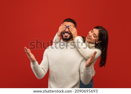 Romantic surprise concept. Cheerful arab woman covering her boyfriend eyes from back, standing behind him, couple posing on red studio background