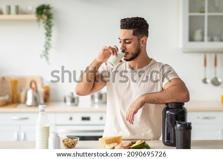Healthy nutrition for muscle gain and weight loss concept. Athletic young Arab man drinking protein shake or milk, standing near table with healthy wholesome products at kitchen, copy space Royalty-Free Stock Photo #2090695726