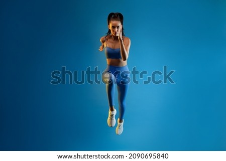 Front-View Of Motivated Sportswoman Running Looking At Camera Over Blue Background. Full Length Shot Of Athletic Lady Runner Exercising In Studio. Sport Motivation Concept