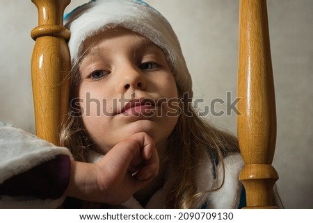 Close-up of a baby girl's face in a snow maiden hat between two balusters on a gray background. Nice child''s face