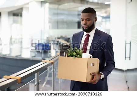 Sad young black man got fired, walking by office building and holding box with his belongings, copy space. Terminated african american guy in suit lost his job, leaving workplace, copy space Royalty-Free Stock Photo #2090710126