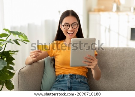 Excited asian woman shopping from home, using digital tablet and credit card, buying things in web store, sitting on sofa, copy space. Sale, retail and purchase concept