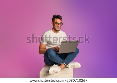 Full length of young Arab man sitting cross legged with laptop, having video call, working online in neon light. Happy middle Eastern guy watching movie, chatting on internet