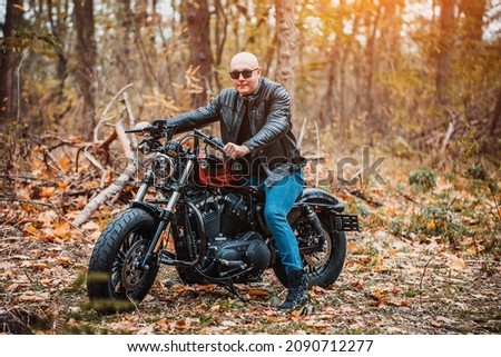 Bald brutal man in sunglasses and a leather jacket sits on a motorcycle on the road in the autumn forest