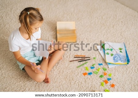 High-angle view of pretty little girl drawing making picture sitting on floor at children room with modern light interior. Childhood and creativity concept.