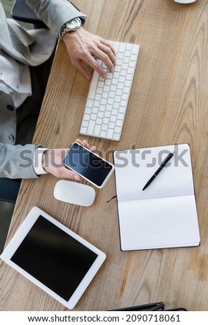 Cropped view of businessman holding cellphone and using computer keyboard in office