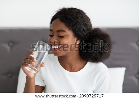 Happy young beautiful african american woman drinking fresh water and smiling, sitting in bed, start new day with fresh aqua, body hydration in the morning concept, closeup photo