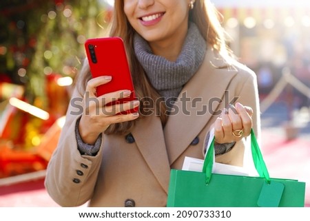 Beautiful woman holding smartphone for shopping online and carrying bags on Christmas time. Cropped picture with focus on the hand and phone.