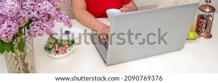 On table are lilacs, laptop, tea, sweets. An older woman's hand is visible behind laptop. Selective focus. Picture for articles about people and technology.