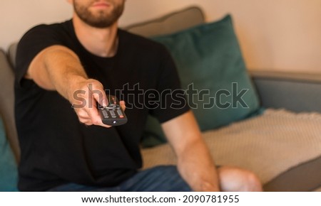 Young bearded man sitting on couch while switching TV channels. Focus on remote control. close up Television remote control in casual man hands pointing to tv set and turning it on or off
