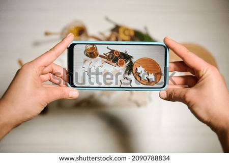 Phone in real time. Hands holding phone and taking pictures of gingerbreads laid out on wooden board and on baking paper sprinkled with sugar powder next to dried orange slices, pine cones and twigs