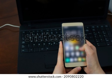Woman's hand holding mobile phone with the laptop in the background. Woman trying to call laptop support or service for broken technical device. Lifestyle
