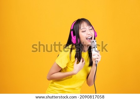 Pretty Asian woman wearing yellow shirt holding microphone singing karaoke music on yellow background. Listen music with headphones sing song in microphone. Royalty-Free Stock Photo #2090817058