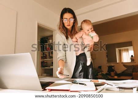 Female interior designer holding her baby while working on a new project. Multitasking mom taking a picture of her notes in her home office. Creative businesswoman balancing work and motherhood.