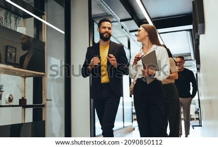 Young professionals having a discussion in a modern office. Two happy young businesspeople smiling while walking together in a hallway. Cheerful colleagues collaborating on a new project. Royalty-Free Stock Photo #2090850112
