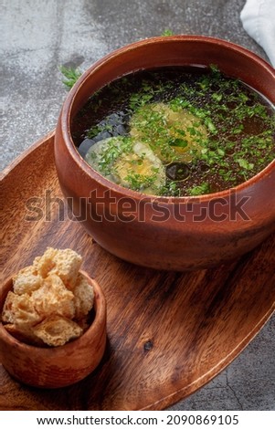 Chicken soup with boiled egg sprinkled with dill and parsley greens in a wooden bowl on a tray with bread crumbs, standing on a stone table background 