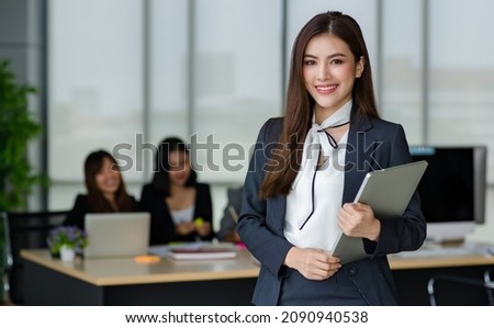 Portrait of young attractive Asian female office worker in formal business suits  smiling at camera in office with blurry colleagues sitting in office as background. Royalty-Free Stock Photo #2090940538