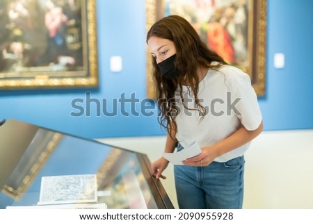Focused young girl in a protective mask, visiting a museum during a pandemic, looks at the exposition behind a glass..case, holding an information booklet Royalty-Free Stock Photo #2090955928