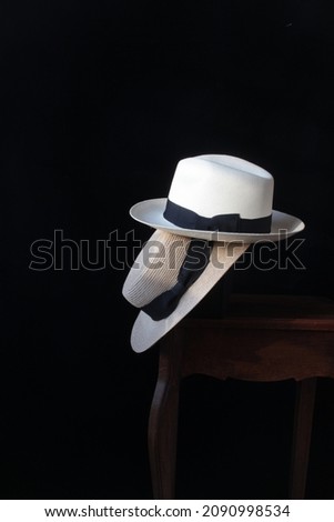 A closeup picture of two hats overlapped on a wooden table: a lady's boater and a gentleman's Panama hat. They were arranged in front of a black background.