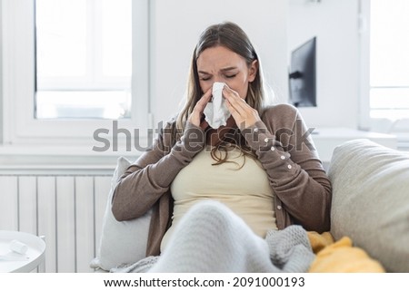 Pregnancy and illness. Sick pregnant woman blowing nose in tissue having fever sitting on sofa indoor suffering from covid-19