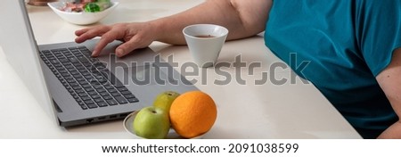 Man's hand in return presses laptop buttons. There is mug of tea nearby. Fruits on saucer are blurred in foreground. Selective focus. Picture for articles about people and technology.