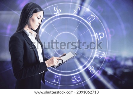 Attractive young european businesswoman with tablet and abstract astrological wheel hologram on blurry office interior background. Business forecasting and future concept. Double exposure