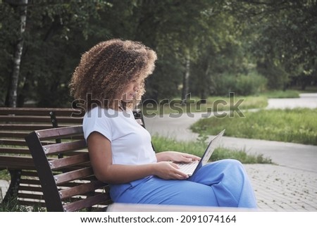 Modern African American woman focused remotely working use laptop sitting on bench at summer park. Female freelancer enjoying distance online communication chatting surfing internet on computer