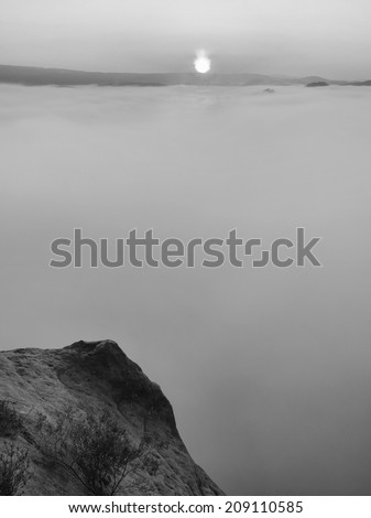 Dense fog in deep valley in Saxony Switzerland full of heavy clouds. Sandstone peaks increased from foggy background, the Sun above. Black and white picture. 