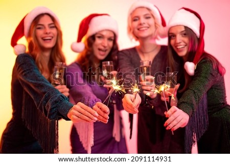 Four smiling happy girls in festive New Year's dresses and Santa's hats holds sparklers and glasses of champagne while celebrating New Year or Christmas on colorful background. Selective focus 