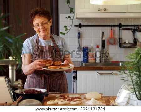 Portrait of an active elderly woman cooking in the kitchen. Grandma has prepared delicious pastries and is holding a plate of whites. Healthy homemade food. Royalty-Free Stock Photo #2091134734
