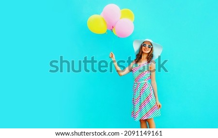 Portrait of beautiful happy smiling young woman with bunch of balloons wearing a colorful dress on blue background