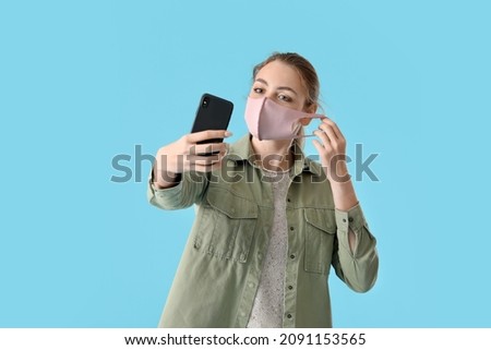 Young woman in medical mask taking selfie on color background