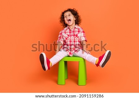 Photo of excited funny school girl dressed checkered clothes sitting green chair looking empty space laughing isolated orange color background