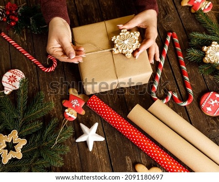 Woman holding a snow flake shaped winter holiday home baked cookie with festive sugar icing above a Christmas gift placed on wooden table background with evergreen branches and other candy treats
