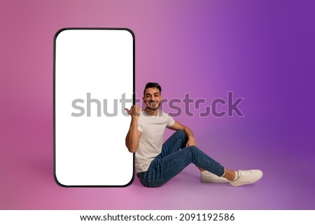Full length of cheerful young Arab man sitting near big cellphone with mockup for mobile app on screen in neon light. Smartphone display template for new website or online ad Royalty-Free Stock Photo #2091192586