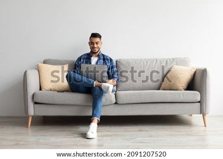 People And Technology. Portrait of young smiling Arab man holding pc on lap sitting on the sofa in living room, typing on keyboard. Cheerful guy browsing internet, surfing web, free copy space Royalty-Free Stock Photo #2091207520