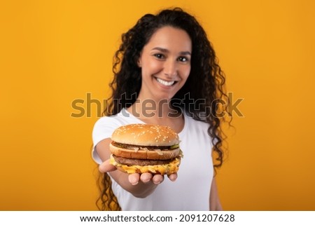 Enjoy Your Meal. Portrait Of Smiling Woman Holding Big Tasty Burger In Hand Showing Sandwish On Palm Close To Camera, Lady Giving And Offering Fastfood, Yellow Orange Studio Background Selective Focus