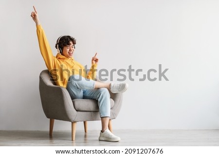 Joyful young Asian guy in wireless headphones sitting in armchair, having fun, listening to music, raising his arm, dancing to favorite song, enjoying cool soundtrack against white wall, empty space Royalty-Free Stock Photo #2091207676
