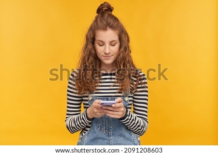 indoor studio shot of young ginger female with long red curly hair keep calm while typing message on her phone isolated over yellow background