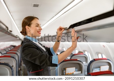 Cheerful flight attendant closing overhead luggage bin in airplane Royalty-Free Stock Photo #2091210307