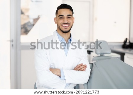 Portrait Of Young Handsome Middle Eastern Dentist Doctor Posing At Workplace In Modern Clinic, Arab Male Stomatologist Wearing Uniform Sitting In Cabinet And Smiling At Camera, Copy Space