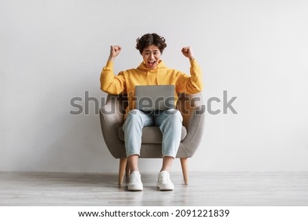Millennial Asian man making YES gesture, sitting in armchair with laptop, celebrating success or achievement against white studio wall. Funky young guy feeling excited over big sale or online win Royalty-Free Stock Photo #2091221839